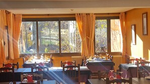A dining area with tables and chairs, large windows with curtains, and a view of trees outside. - Hotel Sucarà (La Cortinada)