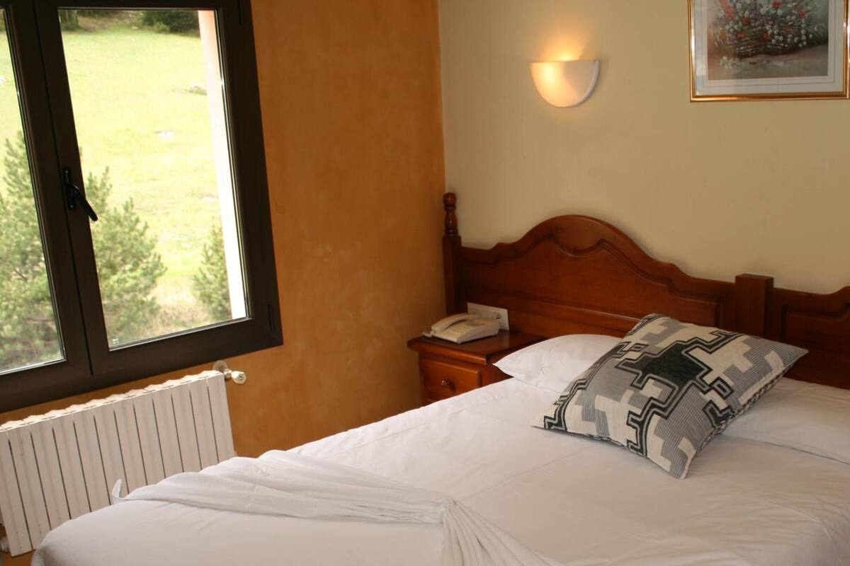 Habitación de hotel con cabecera de madera, una mesita de noche con teléfono, almohada estampada y ventana con vista a follaje.