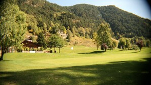 A golf course with a flag, trees, and a house in the background. - Hotel Sucarà (La Cortinada)