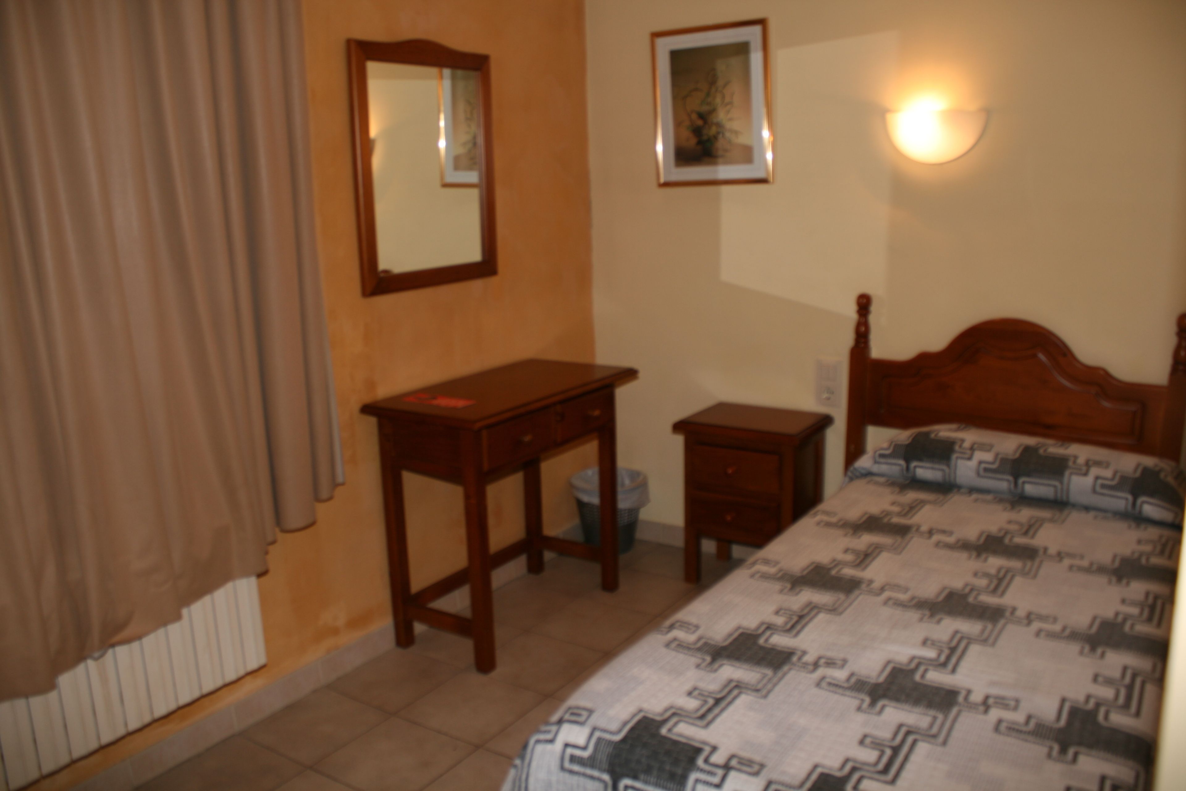 A bedroom with a bed, a wooden nightstand, a desk, a chair, a radiator, a wall-mounted light, and a framed picture.