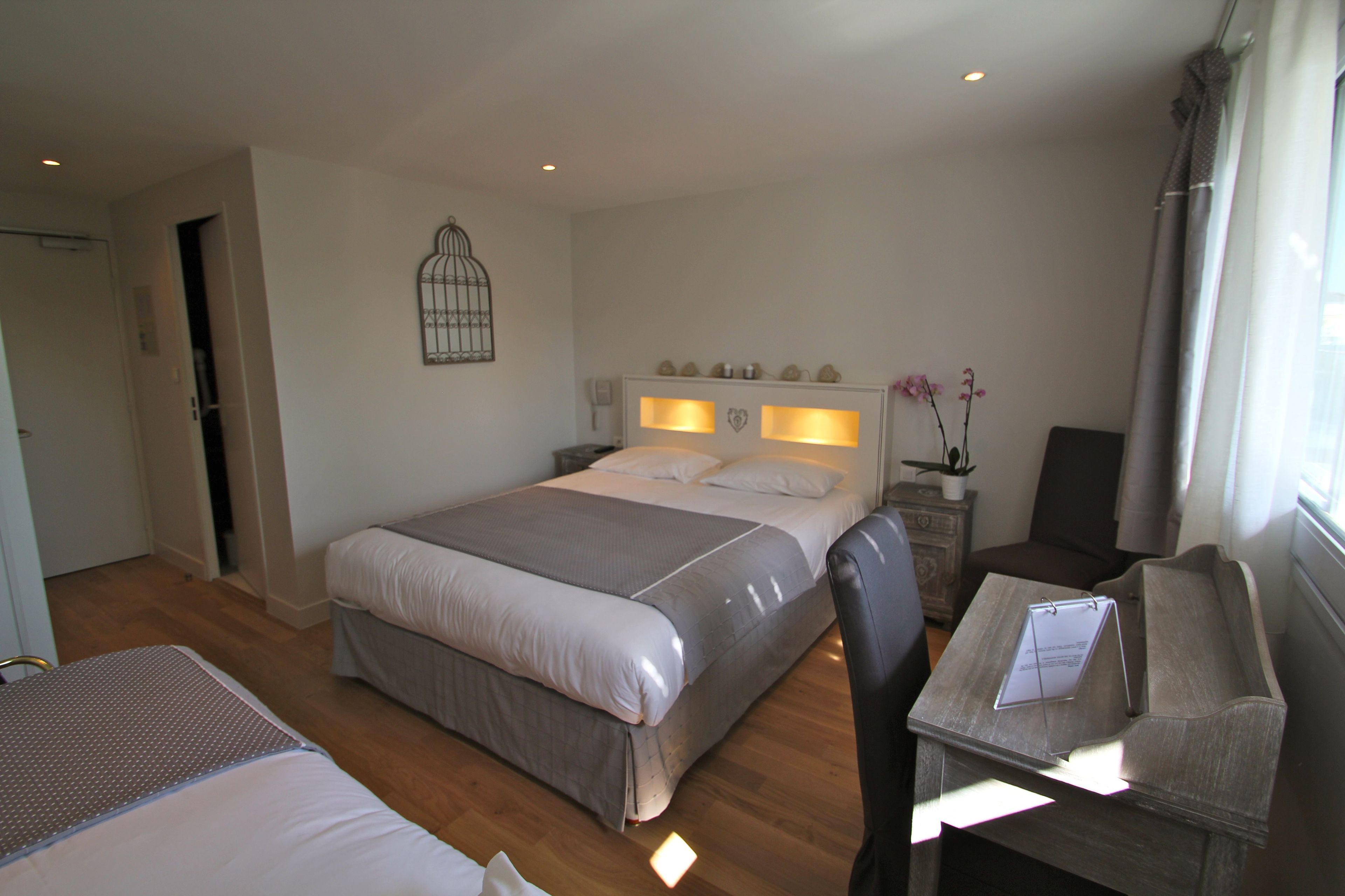 Une chambre d’hôtel comprenant un lit, un bureau avec une chaise, une petite table, une plante en pot et un élément décoratif mural.