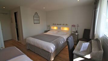Une chambre d’hôtel comprenant un lit, un bureau avec une chaise, une petite table, une plante en pot et un élément décoratif mural.