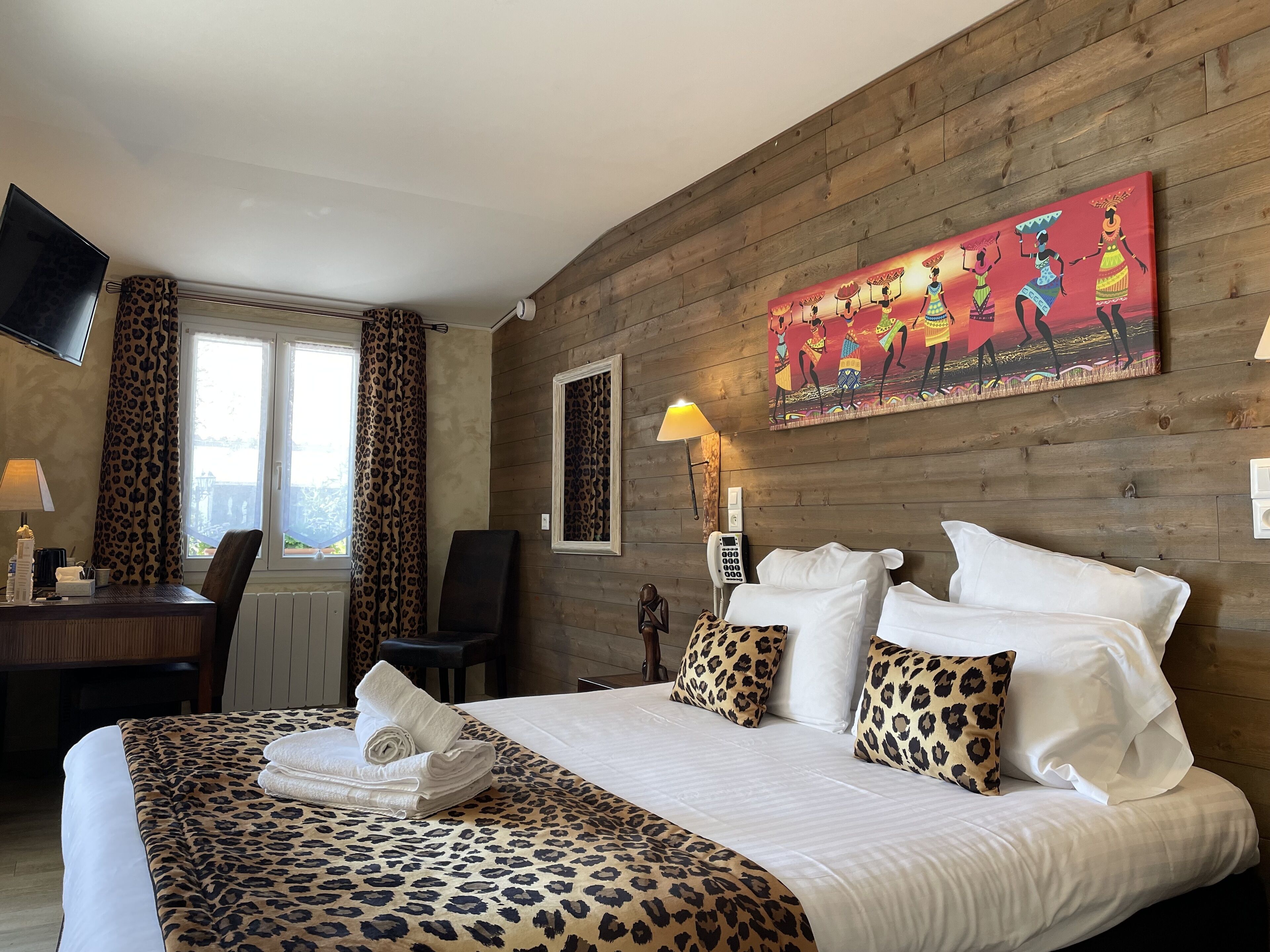 Een hotelkamer met een houten wand, een bed met luipaardprint beddengoed, een bureau met een stoel, een aan de muur bevestigde televisie en een schilderij boven het bed.