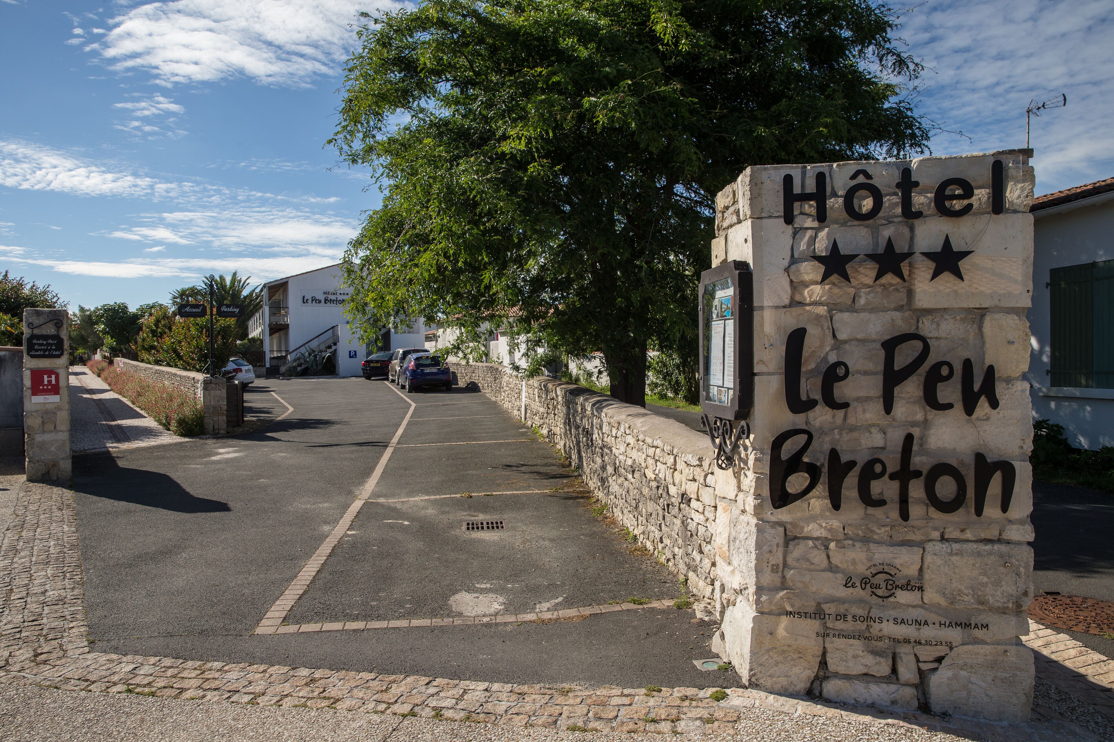 Una señal de piedra indica el Hotel Le Peu Breton, con vista despejada a la calle y coches aparcados.