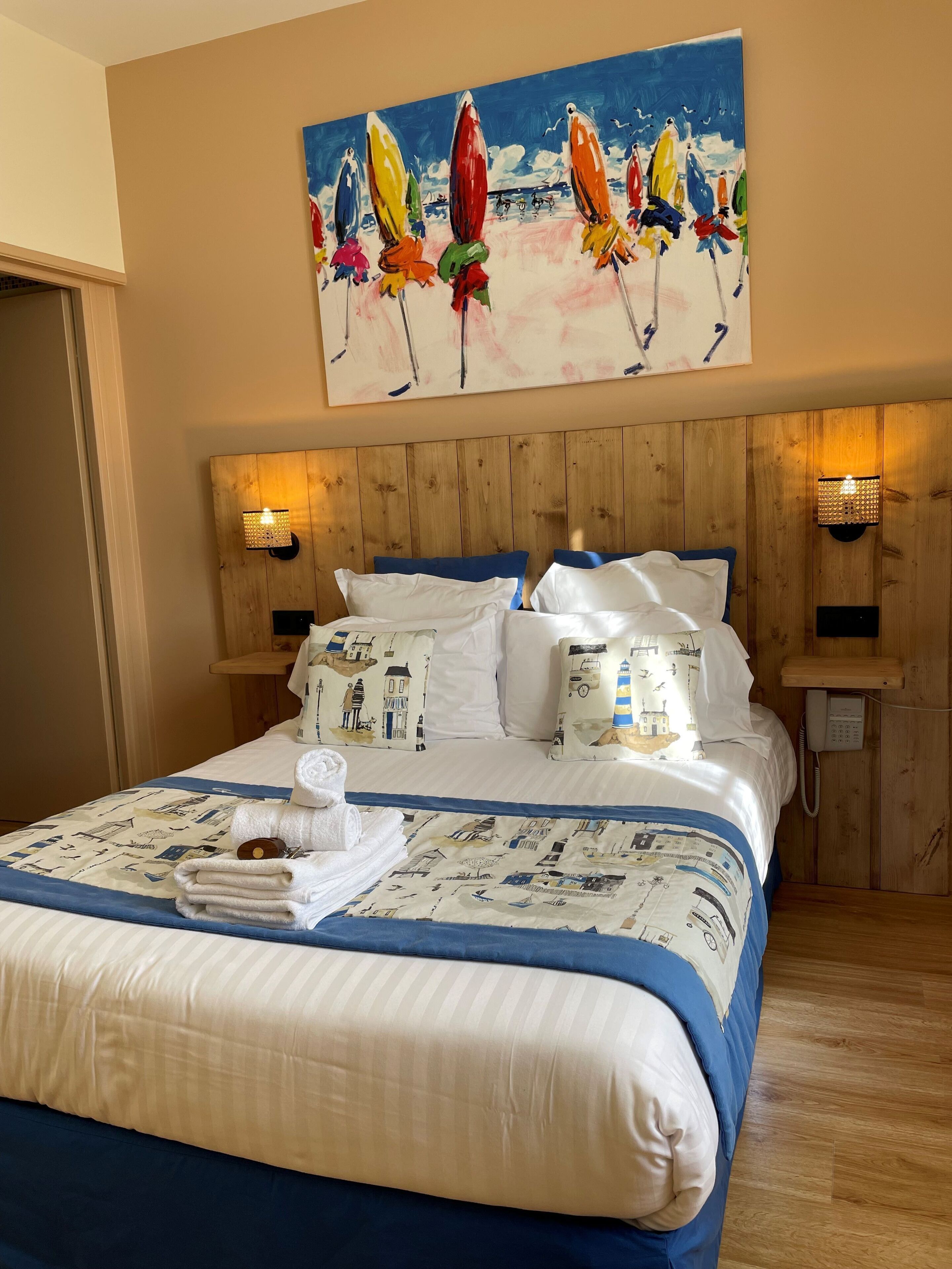 A neatly made bed with a blue and white striped bedspread, white pillows with nautical prints, and a painting of colorful umbrellas above it.