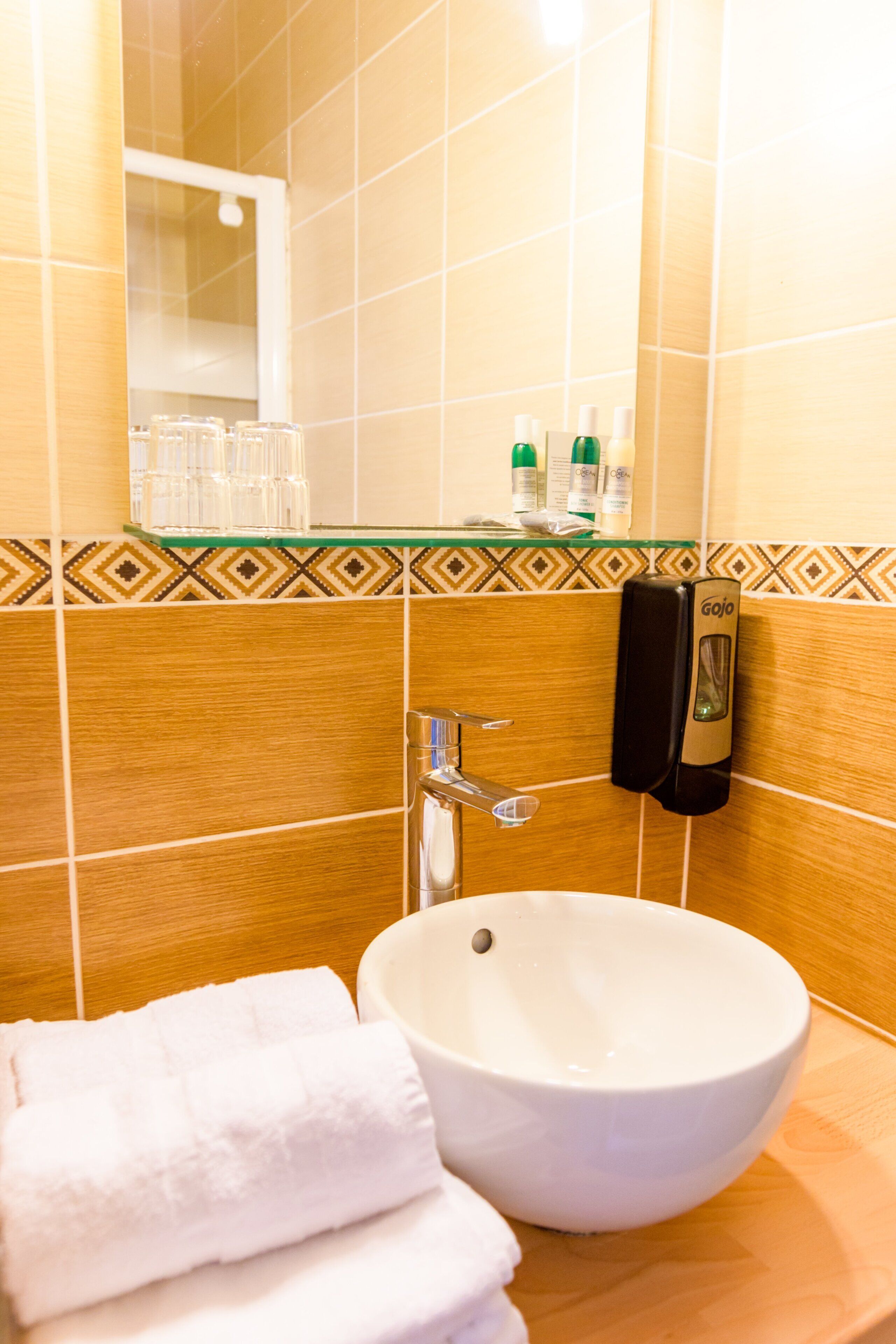 Une salle de bain avec un lavabo blanc, une étagère en verre où sont rangés les articles de toilette et un porte-serviettes.