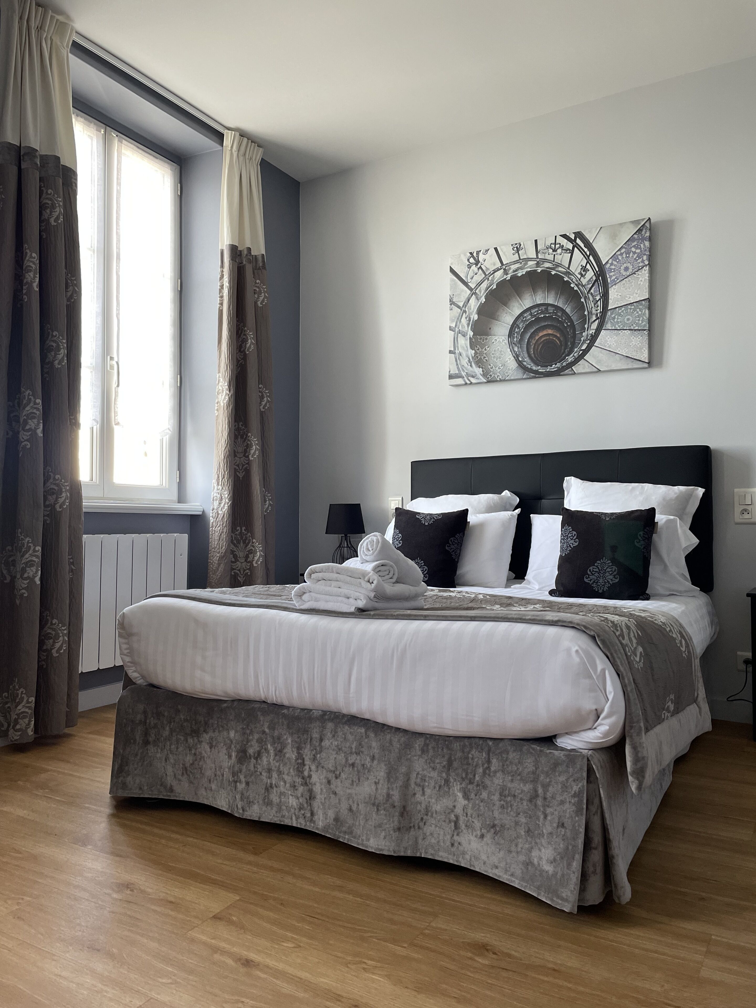 A neatly made bed with white linens and a grey bed skirt, a black headboard, and a wall-mounted abstract art piece.