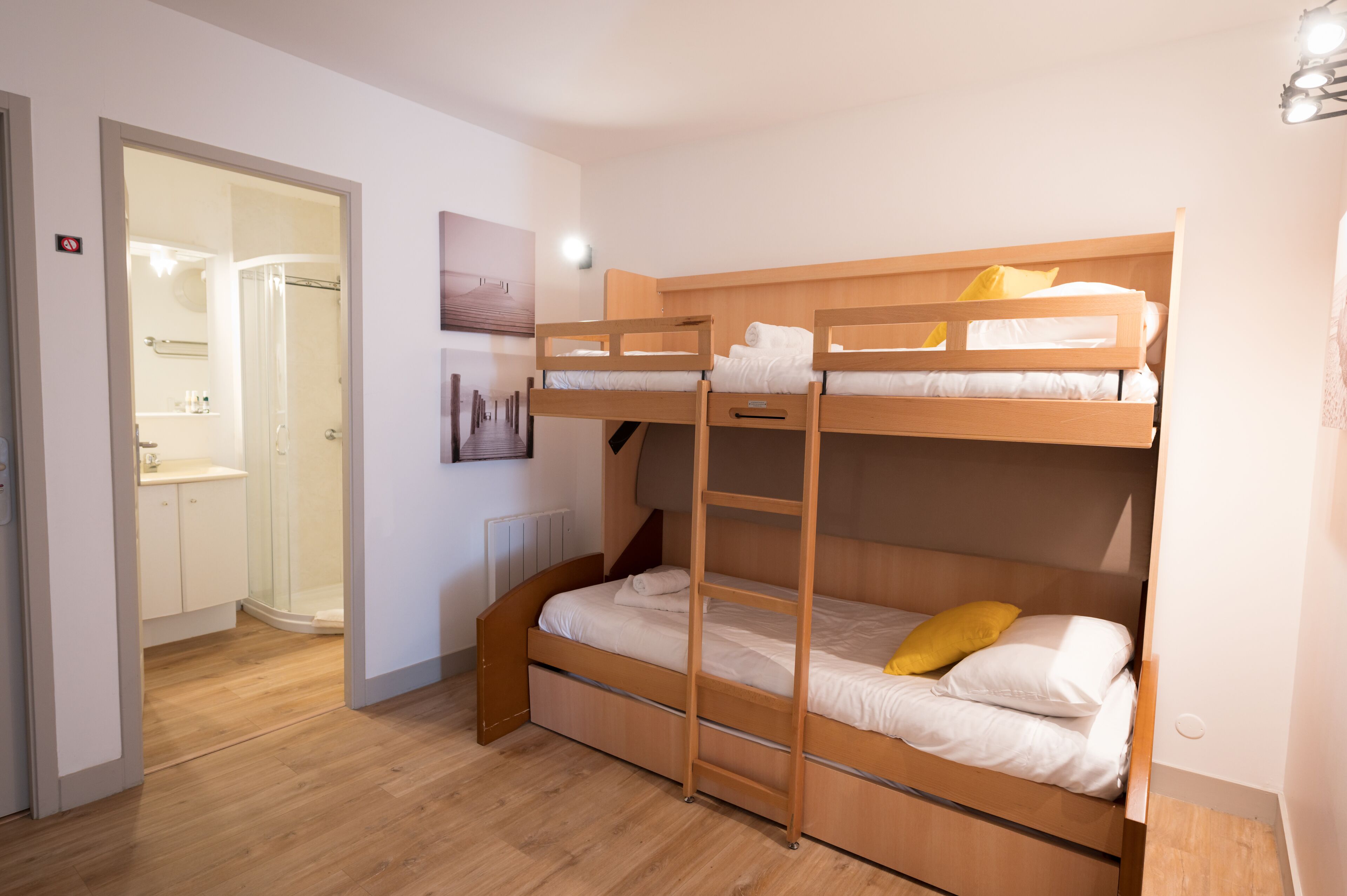 A bunk bed room with a wooden ladder, a bathroom with a glass door, and a white wall with a picture.