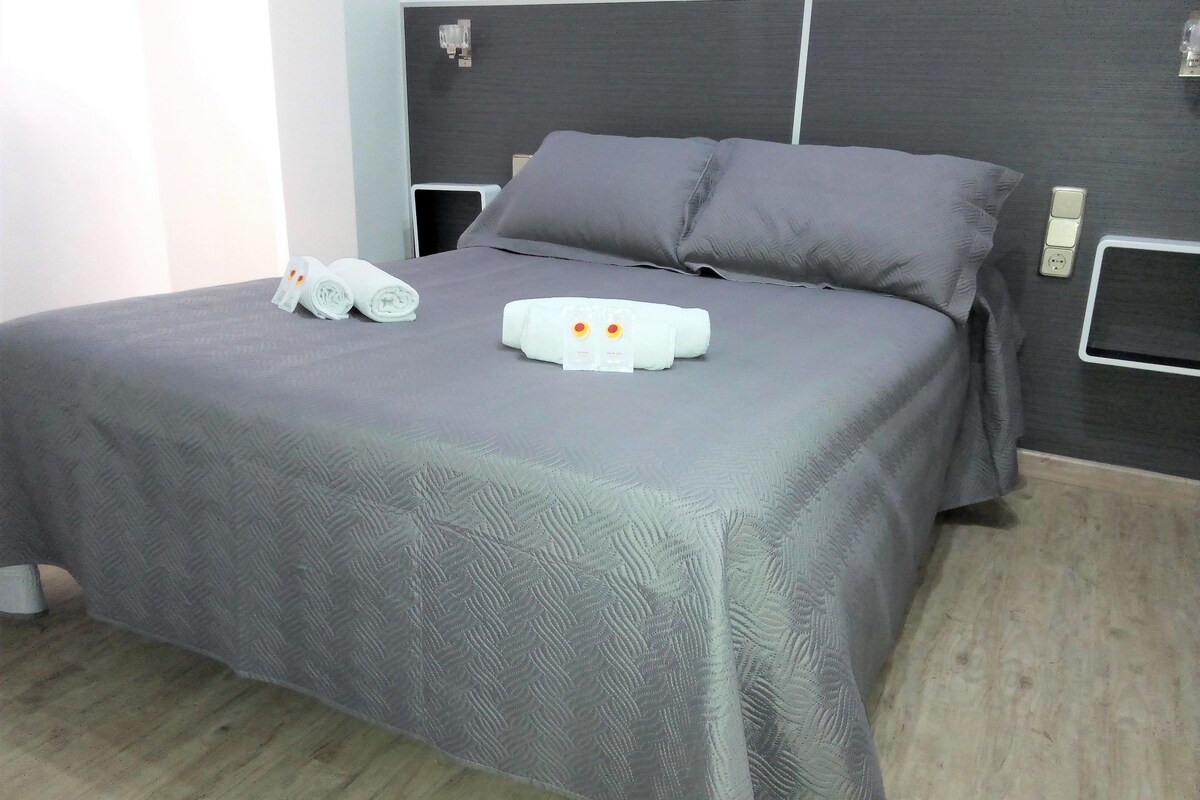 A neatly made bed with a grey textured bedspread, two pillows, and a white toiletry bag with a red tag.