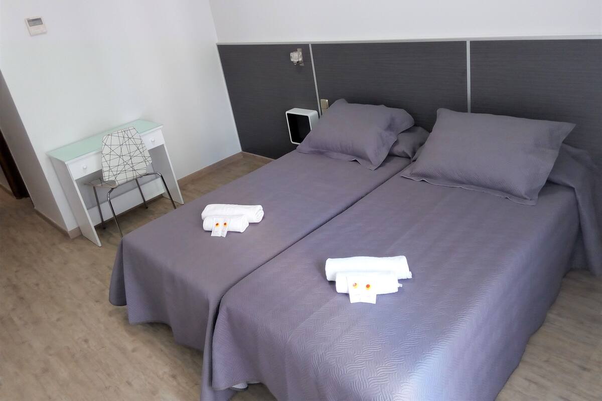 Habitación de hotel con dos camas, cada una con cubrecama morado y toallas blancas. Hay un escritorio con una silla y una mesita con una lámpara.