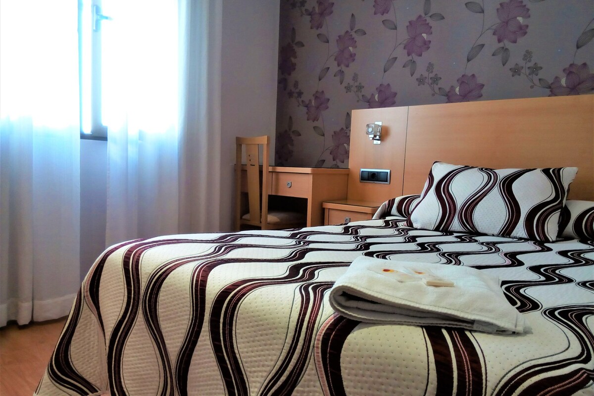 A hotel room with a bed, a wooden nightstand, a desk, and a chair.
