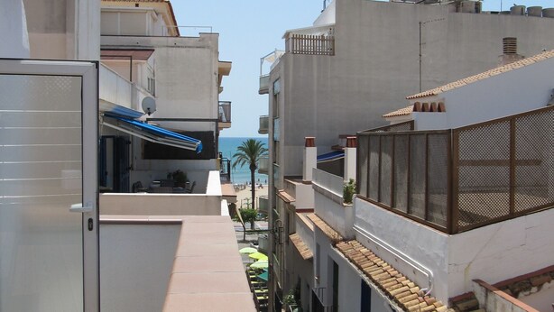 A narrow urban pathway between buildings with balconies, a wooden deck, and a clear view of the sea in the distance. - Hostal Pensió Maricel (Sitges)