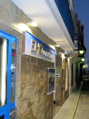 A hotel entrance with a stone facade, a blue door, and illuminated signage. - Hostal Pensió Maricel (Sitges)