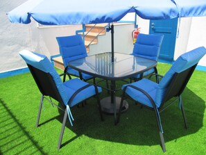 A round glass table with four blue cushioned chairs, an umbrella, and a fire extinguisher in the background. - Hostal Pensió Maricel (Sitges)