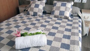 A neatly made bed with a checkered bedspread, pillows, and a rolled towel with a flower. - Hostal Pensió Maricel (Sitges)