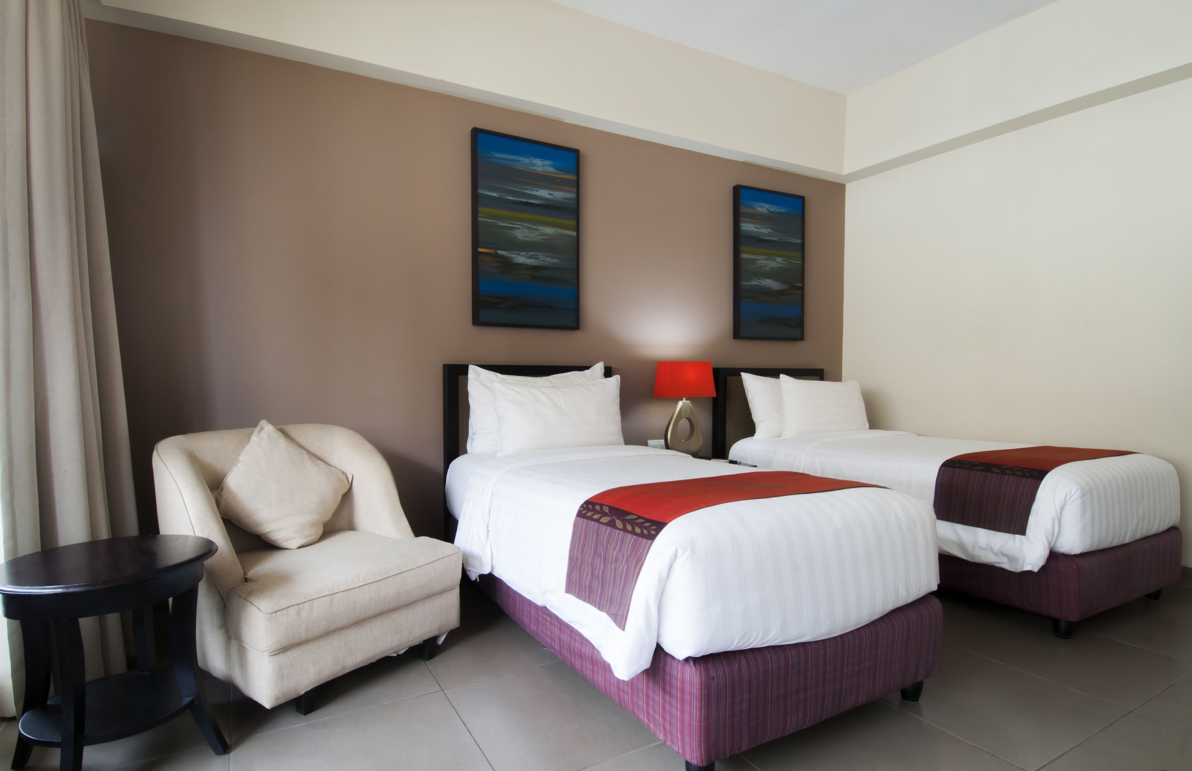 A hotel room with two beds, a chair, a side table, and two paintings on the wall.