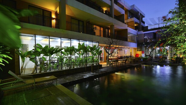 Un hotel de varios pisos con piscina, mobiliario exterior y abundante vegetación.