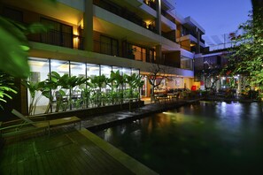 Un hotel de varios pisos con piscina, mobiliario exterior y abundante vegetación.