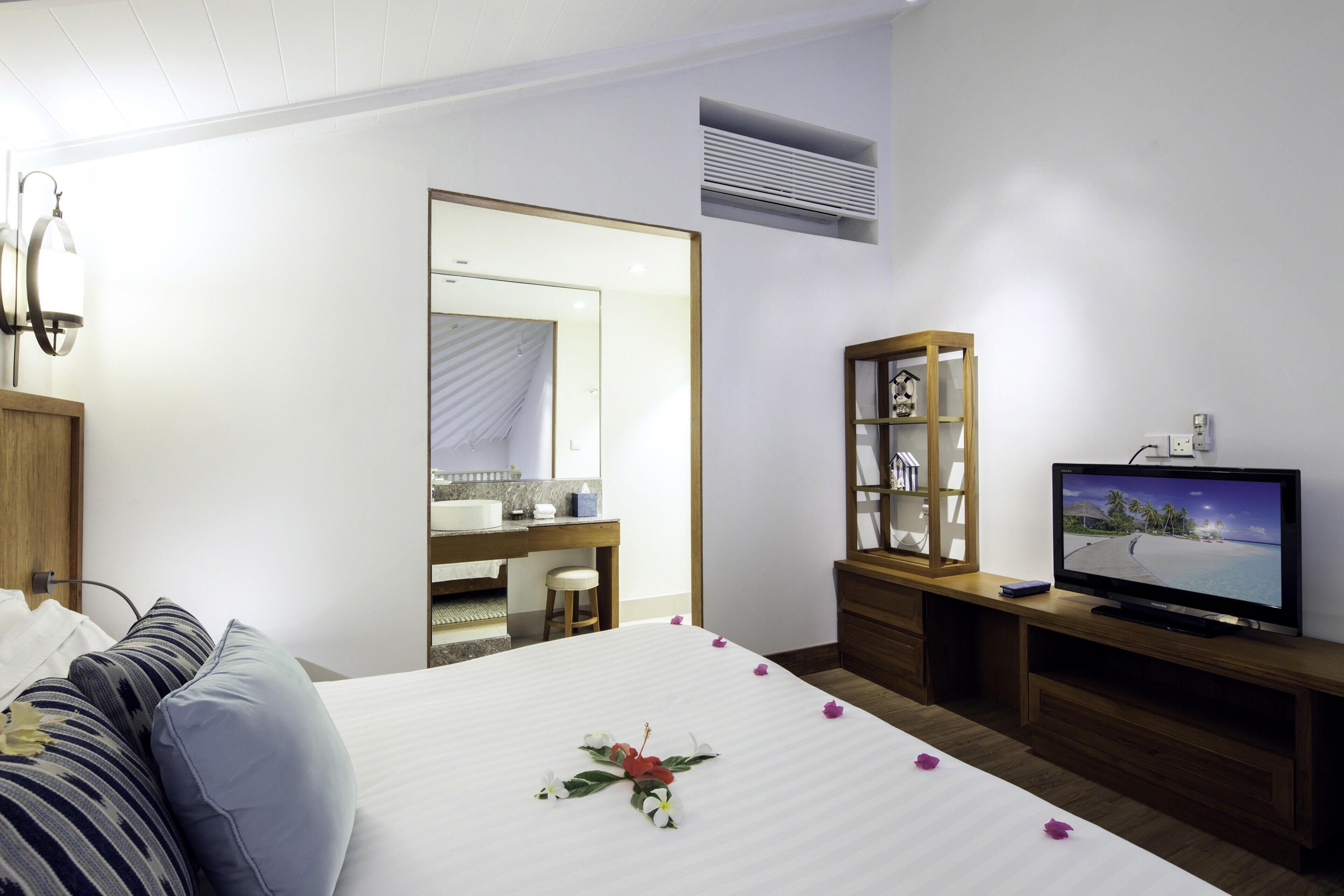 A hotel room with a bed, a television, a wooden cabinet, a mirror, and a bathroom visible through an open door.