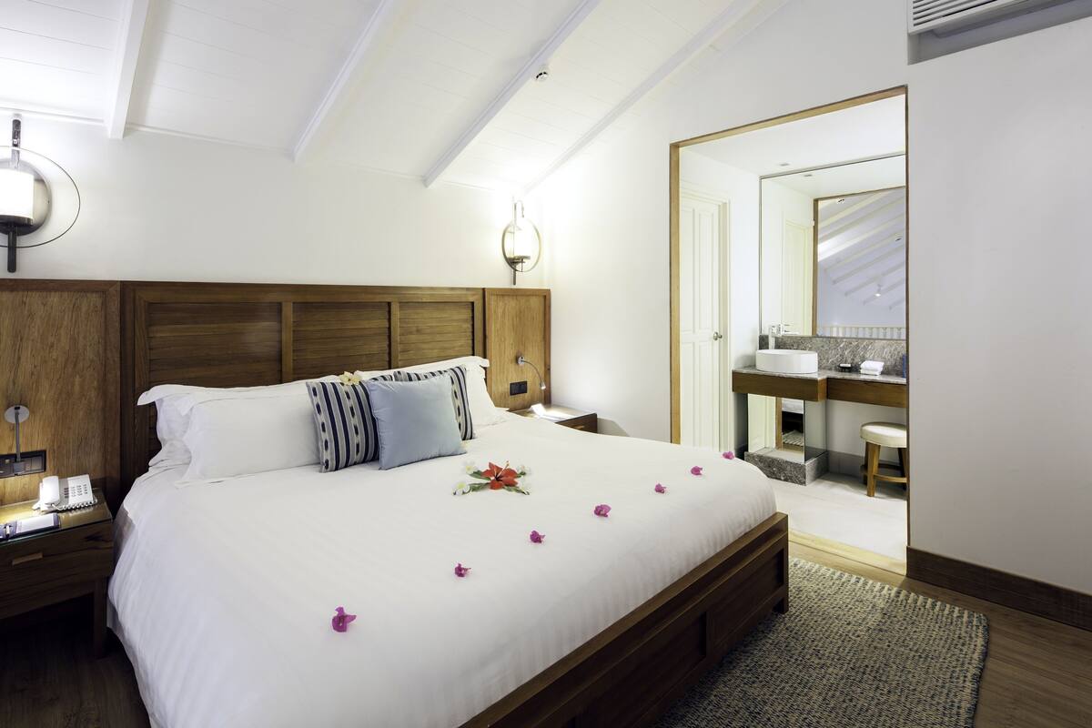 Hotelkamer met een groot bed, nachtkastje met telefoon, houten hoofdeinde, badkamer met spiegel en wastafel, en een deur naar een andere kamer.