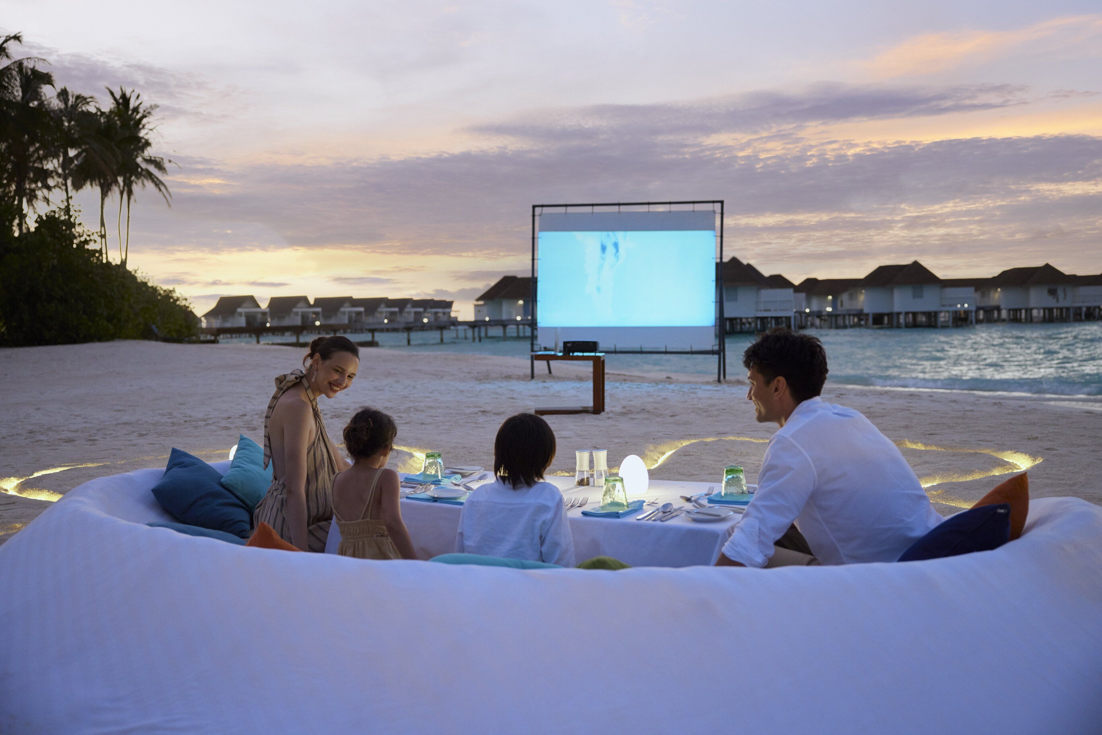 A group of people seated on a white inflatable lounge by the beach with a large screen displaying a video.