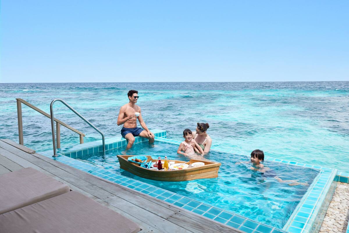 Een zwembadgedeelte met een houten boot vol eten en drinken, een houten terras met ligstoelen en een helder uitzicht op de oceaan.