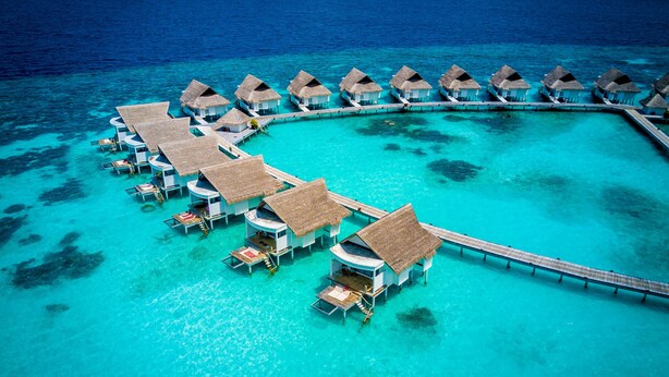 An aerial view of overwater bungalows with thatched roofs, connected by a wooden walkway, in a clear turquoise sea.