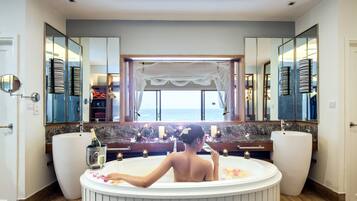 Una mujer en una bañera con vista al mar a través de una ventana, un espejo grande y una botella de champán.