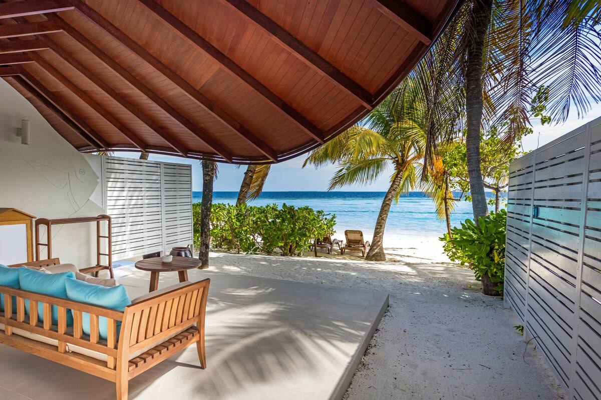 Een overdekt terras met een houten bank en tafel, met uitzicht op een strand met palmbomen en de oceaan.