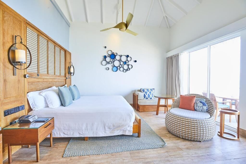 A bedroom with a bed, bedside table, lamp, wall art, and a seating area with a chair and table.