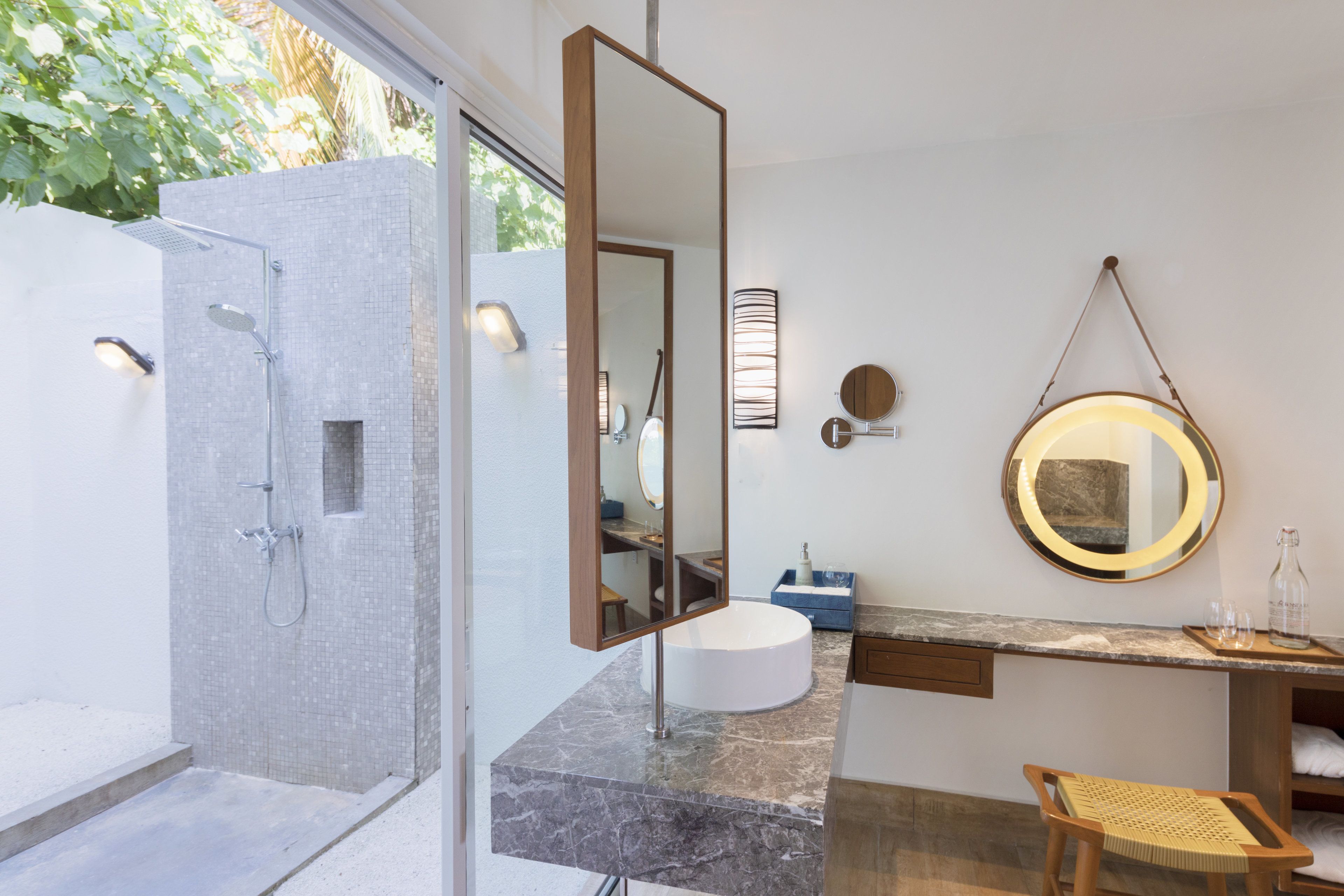 A modern bathroom with a glass-enclosed shower, a white sink, a round mirror, and a wooden chair.