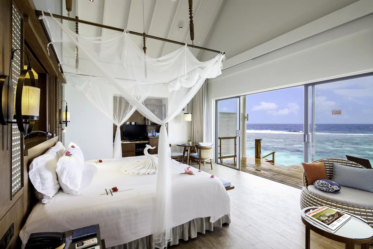 Een luxueuze slaapkamer met een hemelbed, een balkon met uitzicht op de oceaan en een zithoek met een tijdschrift.