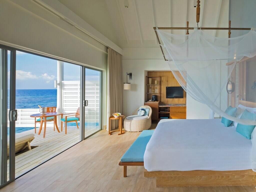 A bedroom with a bed, a TV, a bedside table, a chair, and a view of the ocean.
