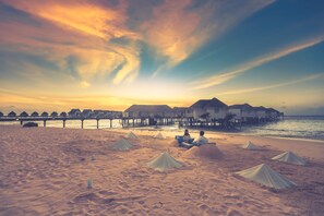 Una playa con bungalows sobre el agua, un muelle y dunas de arena.