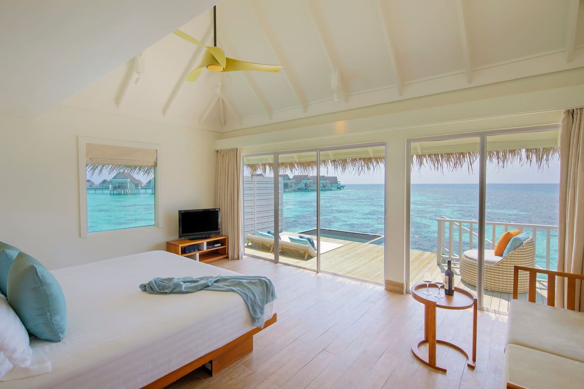 Een slaapkamer met een bed, een televisie, een klein tafeltje met een fles en glas, en een balkon met uitzicht op zee.