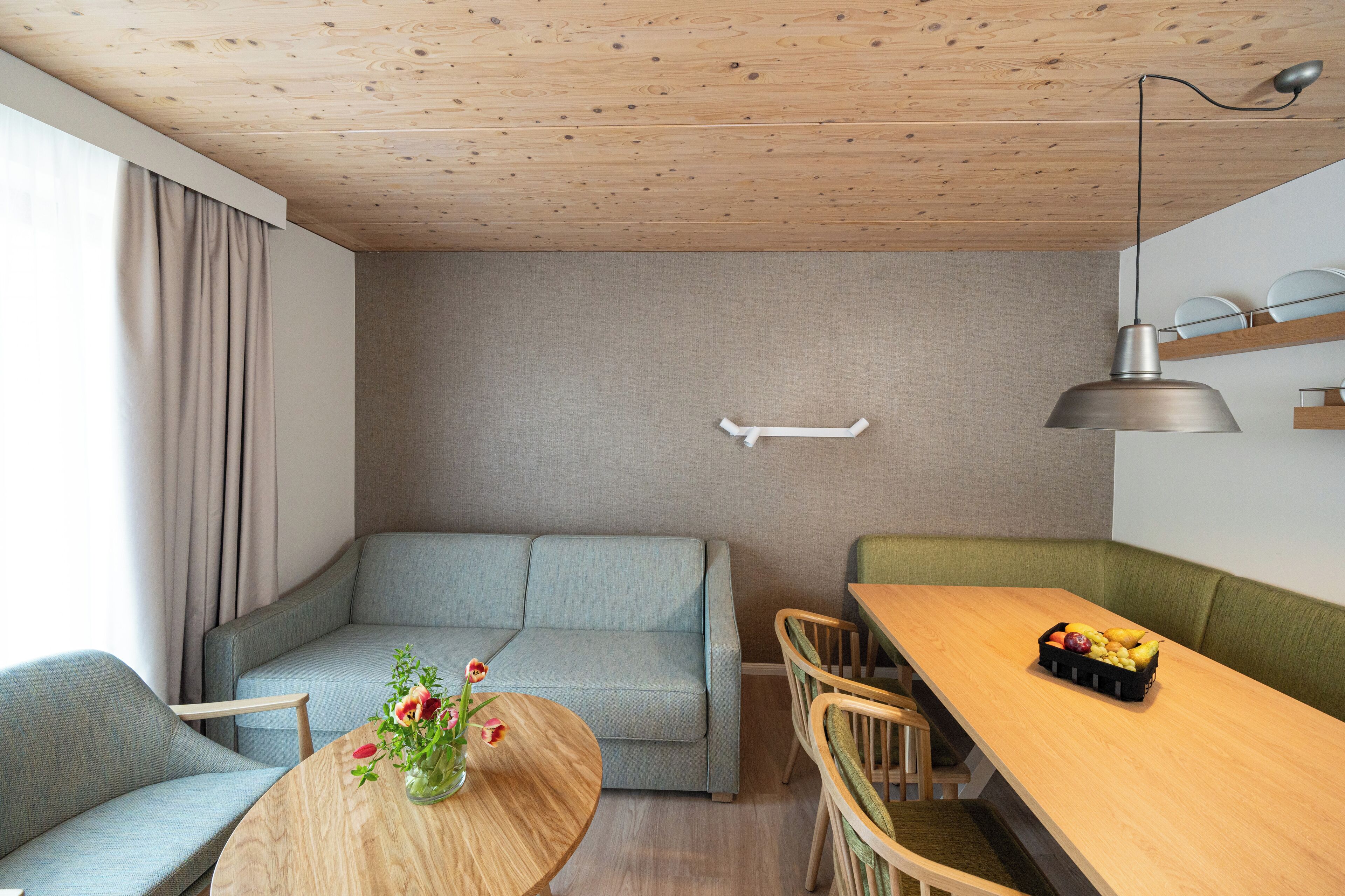 A modern living room with a wooden ceiling, a grey sofa, a round wooden table with a vase of flowers, and a dining area with a wooden table and chairs.