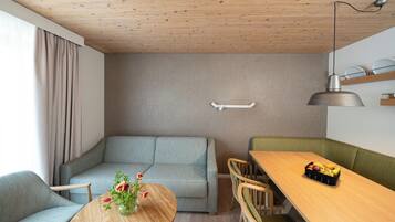 A modern living room with a wooden ceiling, a grey sofa, a round wooden table with a vase of flowers, and a dining area with a wooden table and chairs.