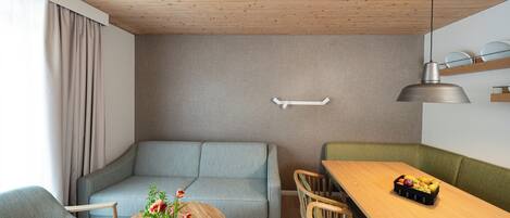 Un salon moderne avec un plafond en bois, un canapé gris, une table ronde en bois agrémentée d’un vase de fleurs et un coin repas avec une table et des chaises en bois.
