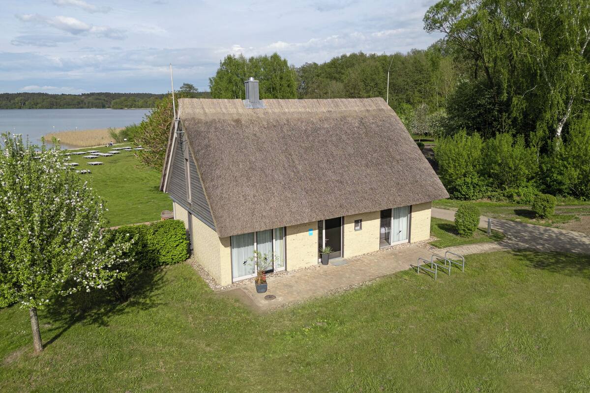 Une maison à toit de chaume, avec une allée pavée, entourée d’une pelouse verdoyante et d’arbres.