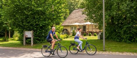 Two people riding bicycles on a paved path with a house and lush greenery in the background.