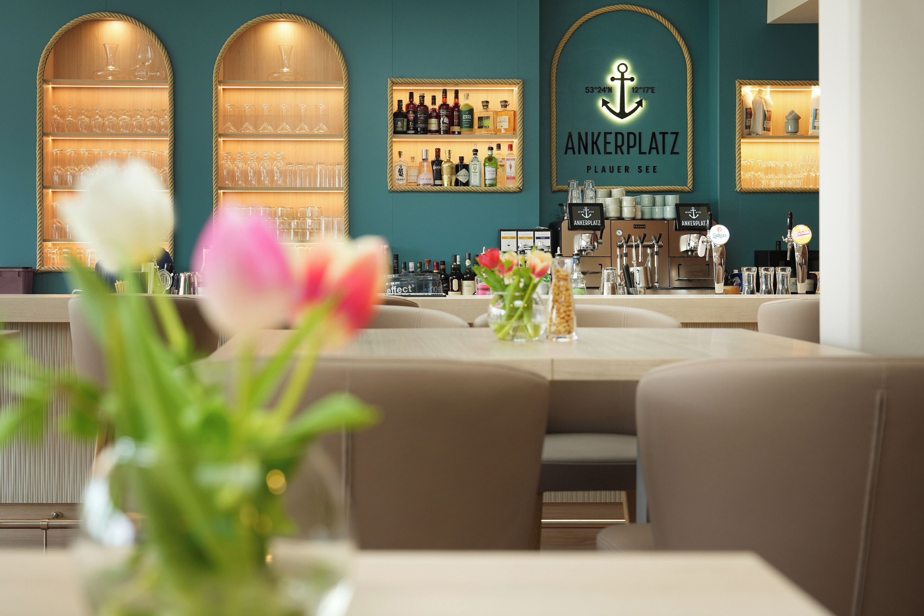 Eine Bar mit Regalen voller Flaschen und Gläsern, einem Schild mit der Aufschrift „ANKERPLATZ“ und einer Vase mit Blumen auf dem Tisch.
