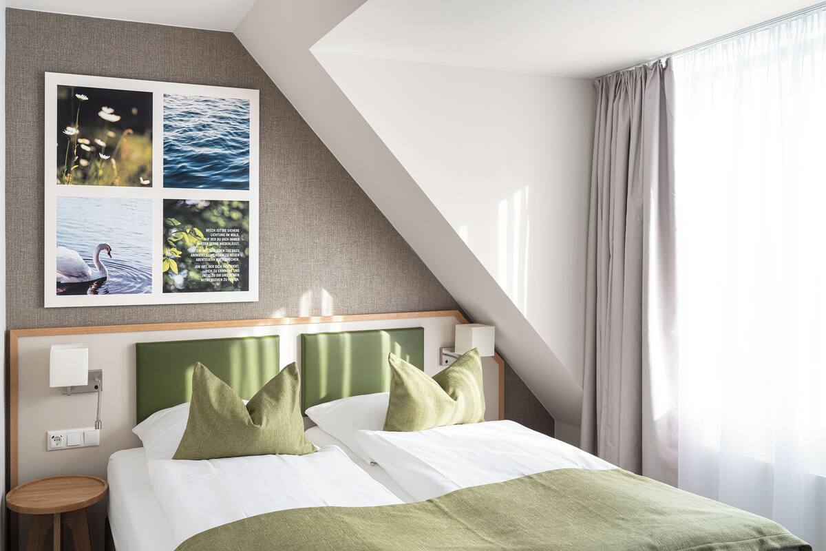 Une chambre d’hôtel moderne, équipée d’un lit, d’oreillers verts, d’une table de chevet, d’une lampe fixée au mur et d’un tableau représentant des paysages naturels.