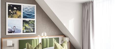 A modern hotel room with a bed, green pillows, a nightstand, a wall-mounted lamp, and a framed picture of nature scenes.