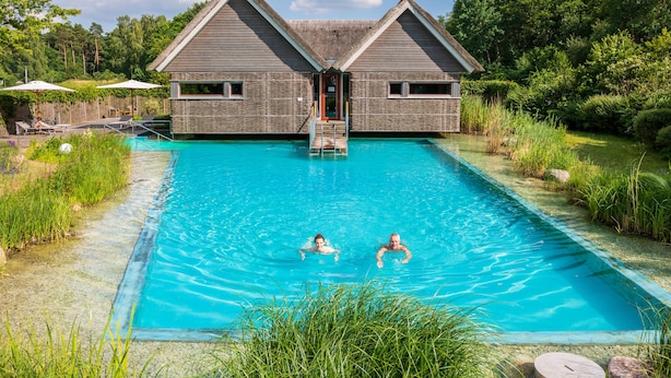 Una piscina amb dues persones nedant, envoltada de vegetació exuberant i una casa de fusta amb teulada de palla.