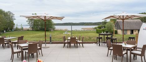 An outdoor patio with tables and chairs, overlooking a lake and a grassy area with a building in the background.