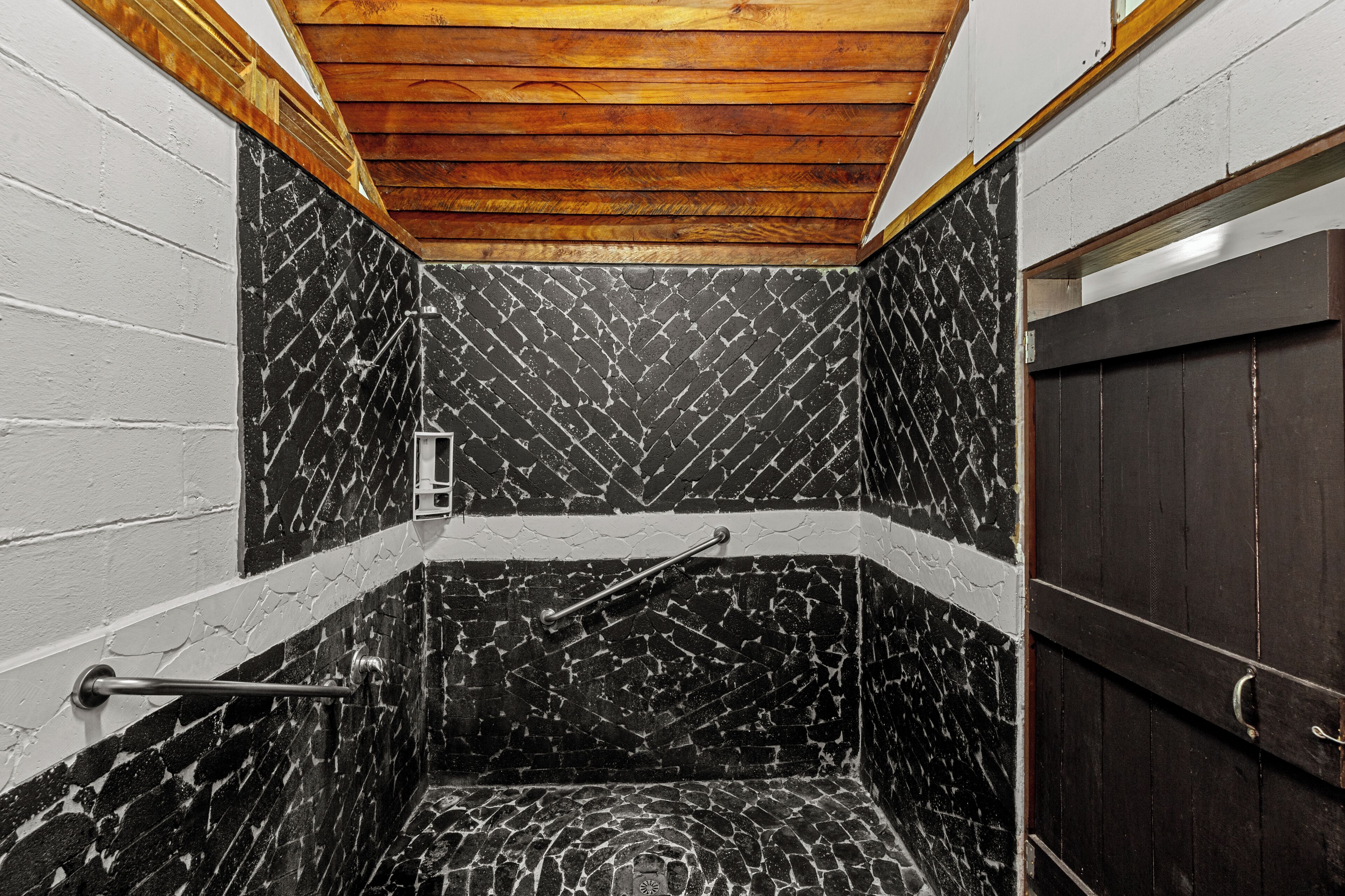 A small, enclosed shower area with black and white patterned tiles, wooden ceiling, and a dark door.