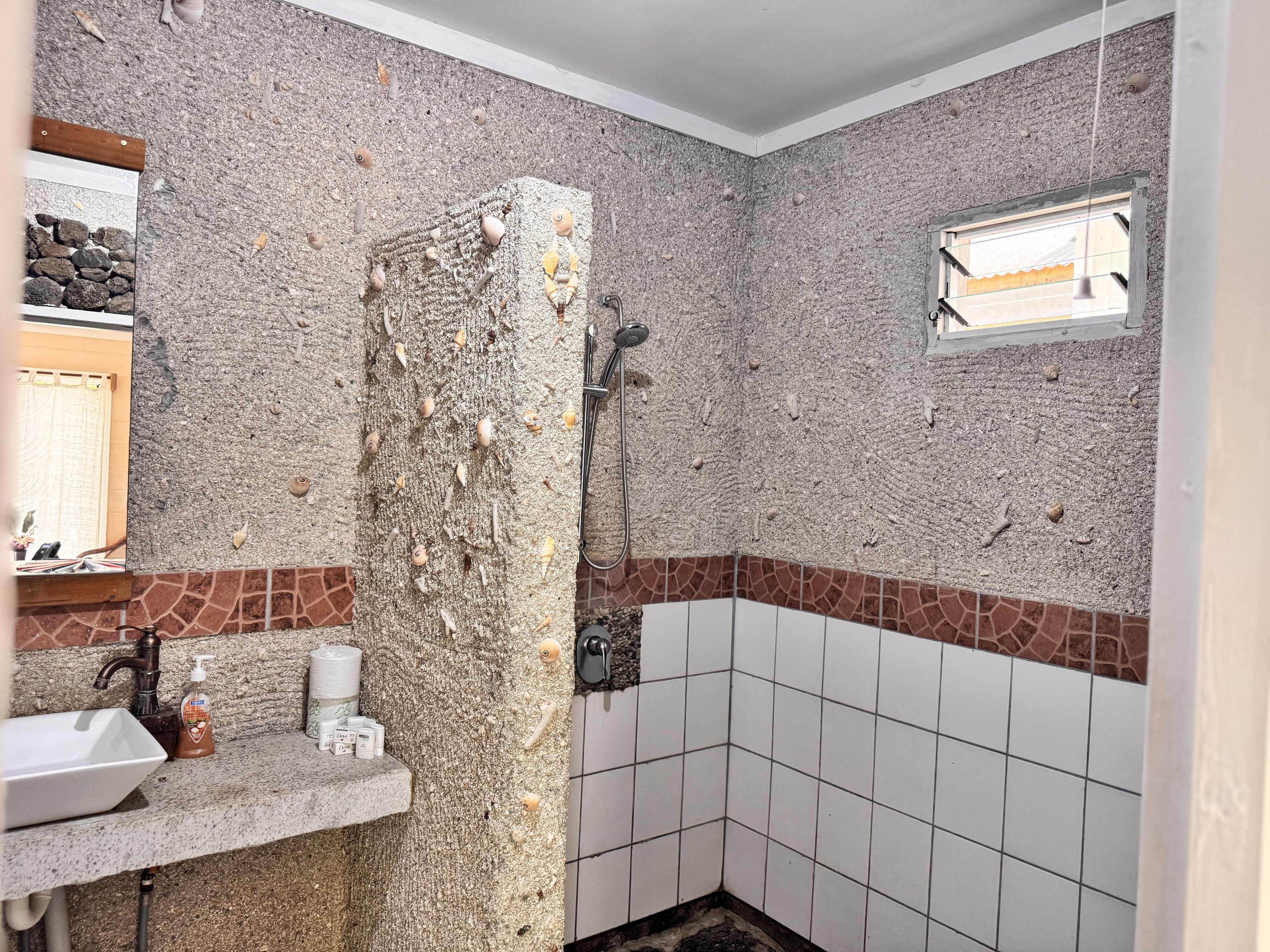 Un baño con pared de textura de guijarros, ducha, lavamanos con artículos de aseo y ventana con vistas.