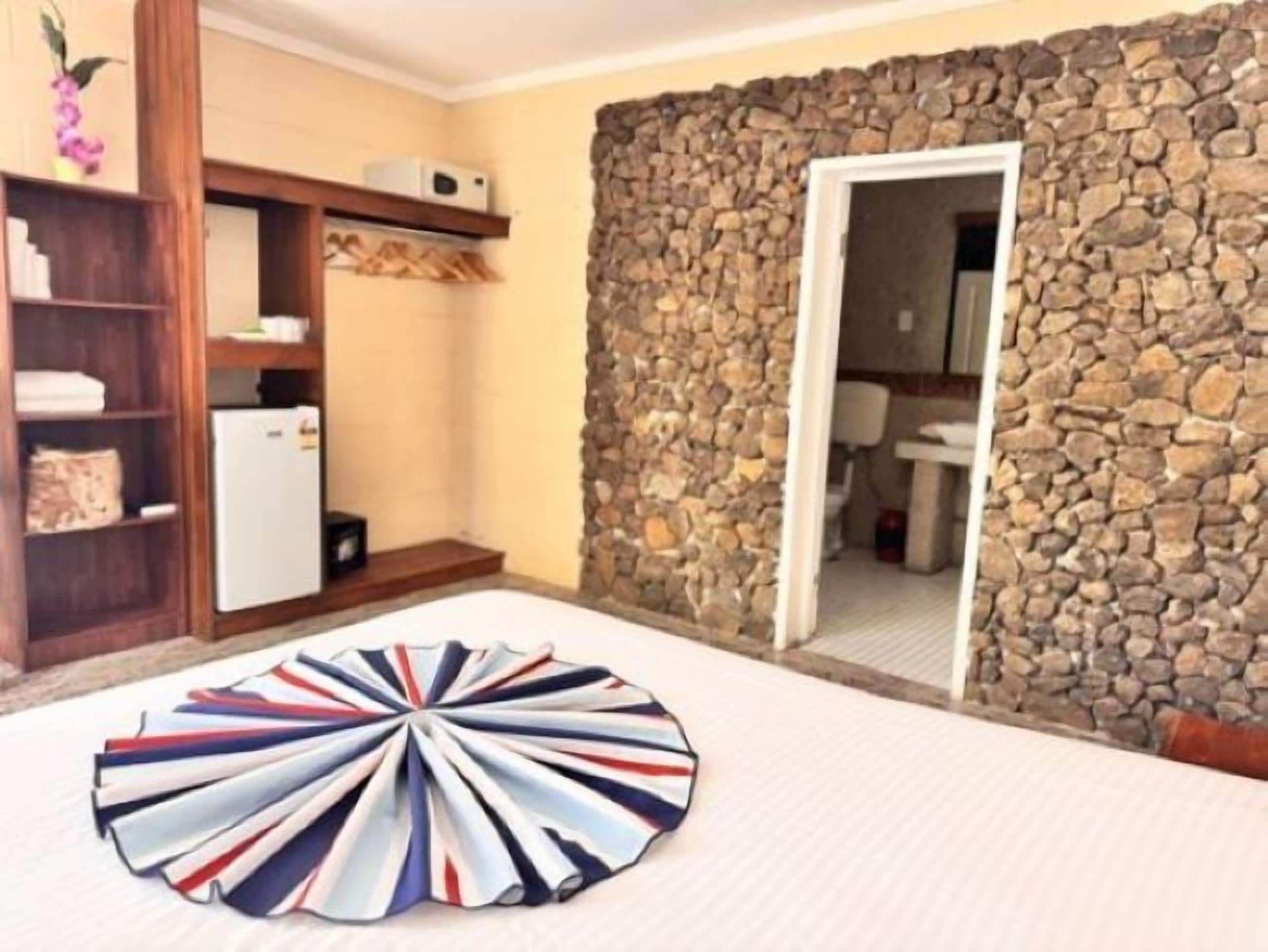 A room with a stone wall, a wooden shelf with towels, a white refrigerator, and a colorful umbrella on the floor.