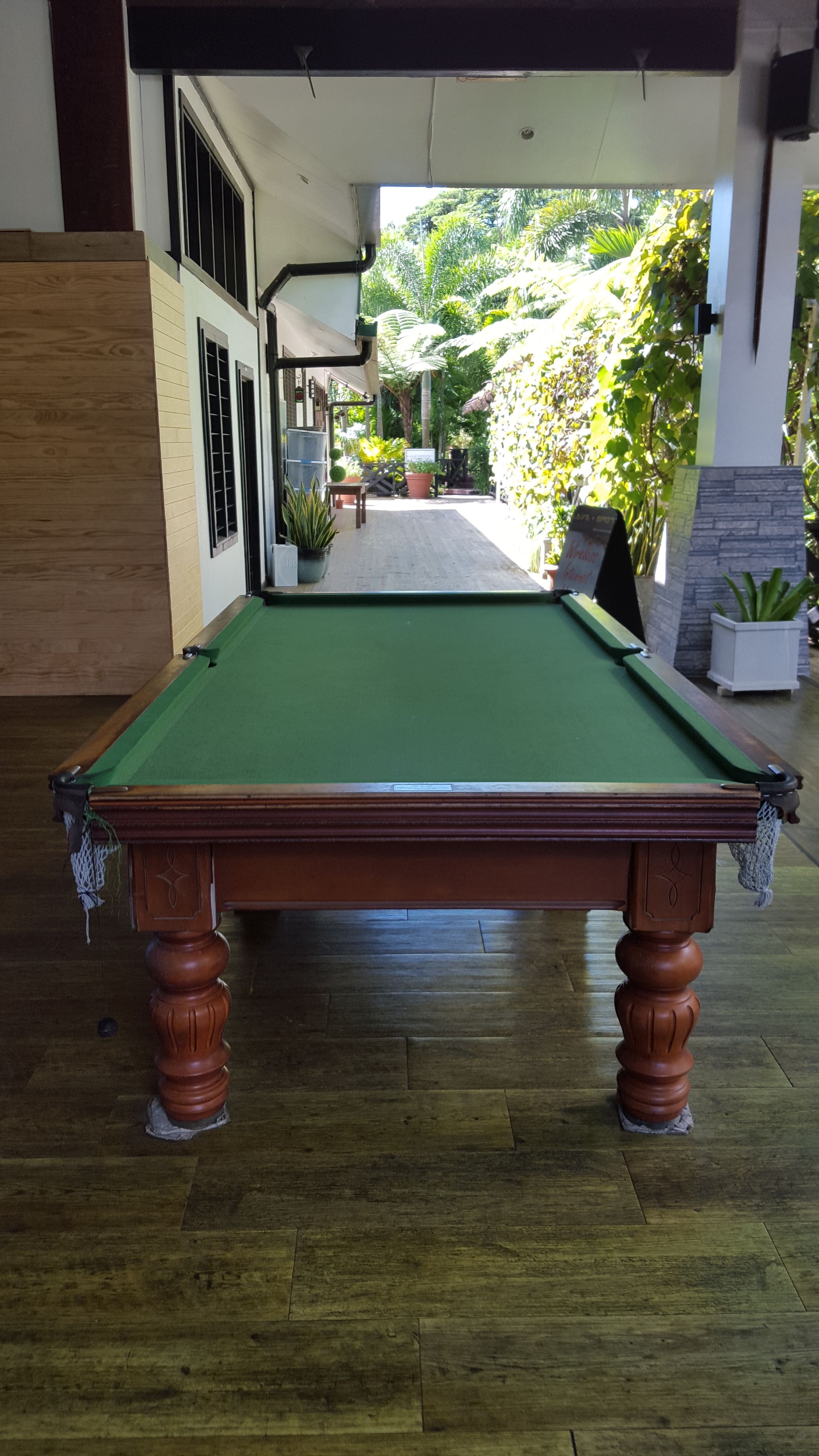 A pool table in a room with wooden flooring, a view of a garden, and a door leading outside.