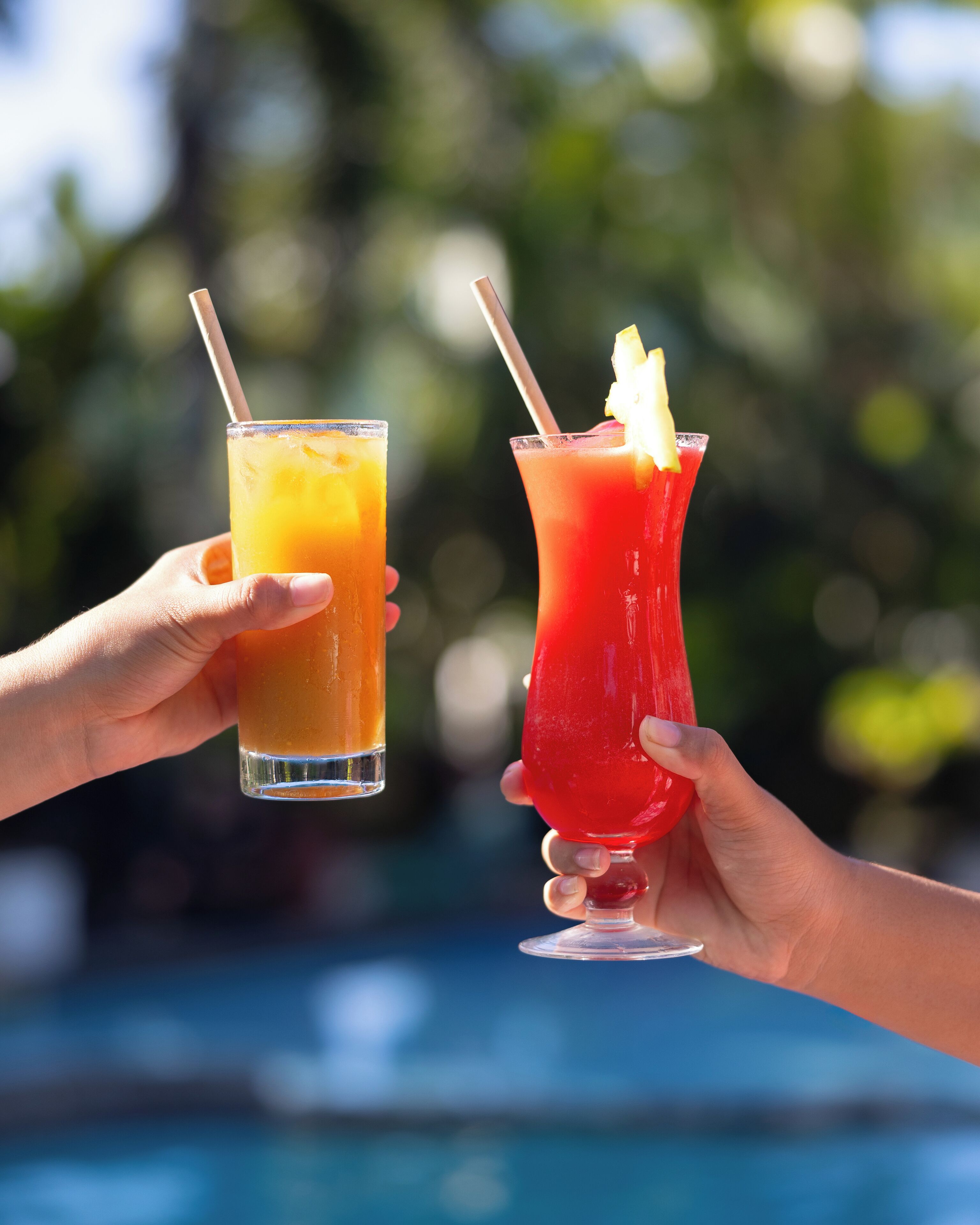 Two hands holding glasses of drinks, one orange and one red, with straws, against a blurred outdoor background.